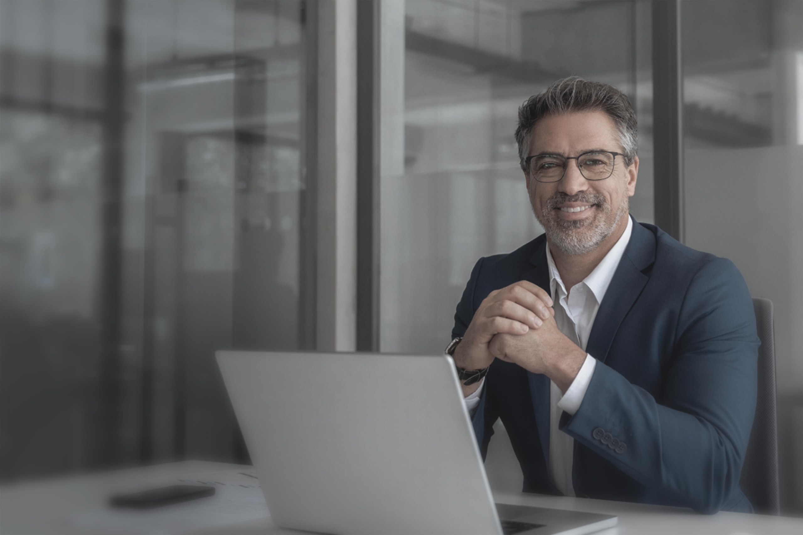 A man sitting in front of a laptop, in a suit, smiling