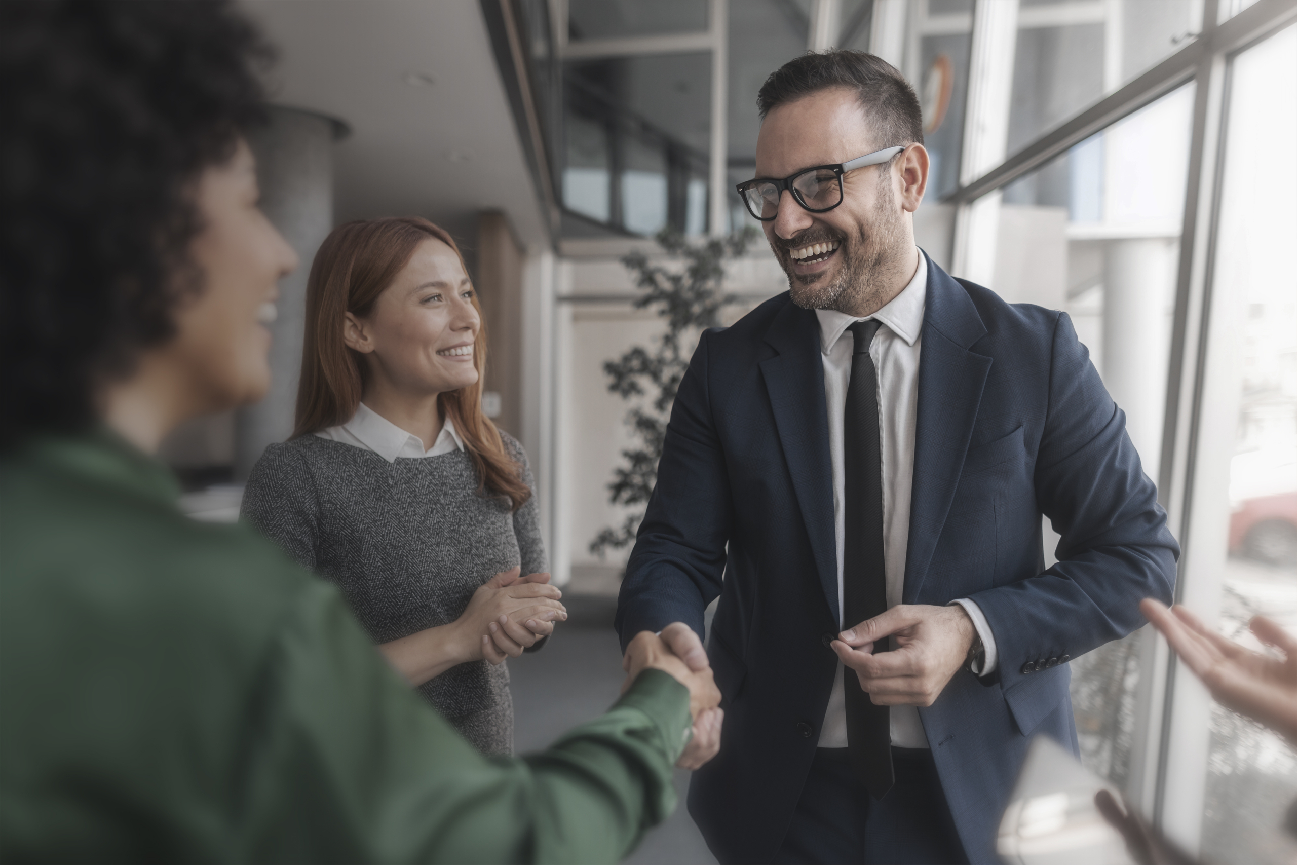 Multiple people in an office environment, a man in a suit, smiling shaking hands with someone