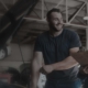 Smiling mechanic talks to a colleague holding a clipboard while working under the open hood of a car in a workshop.